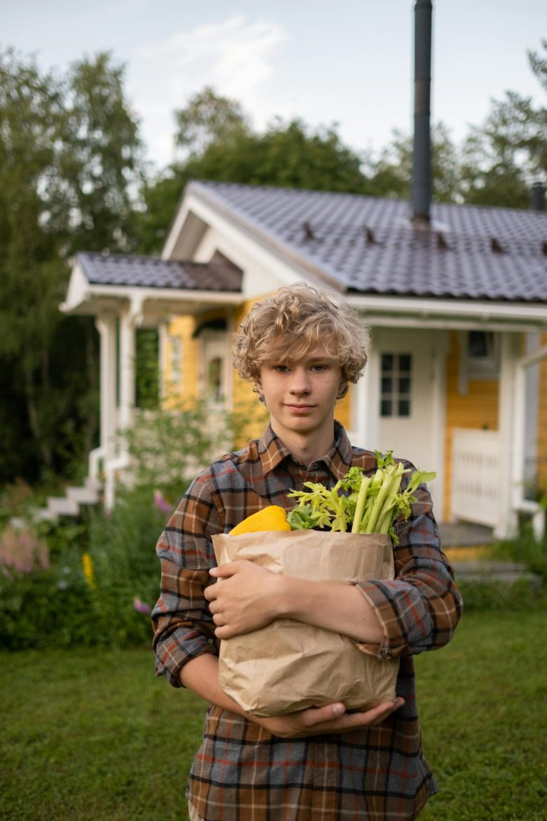 Teenager with curly hair holds a paper bag of groceries in a serene suburban backyard setting.