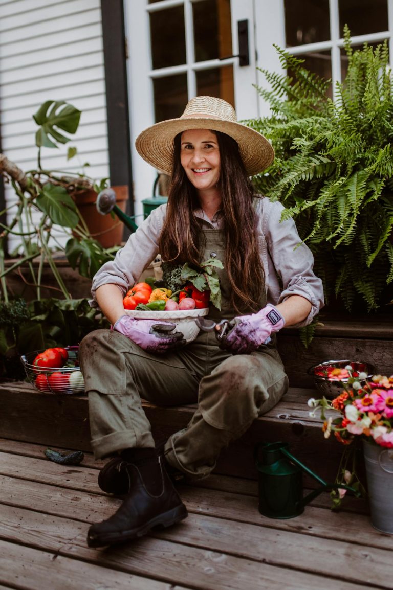 Woman gardener with long hair and straw hat holding a fresh harvest of vegetables.